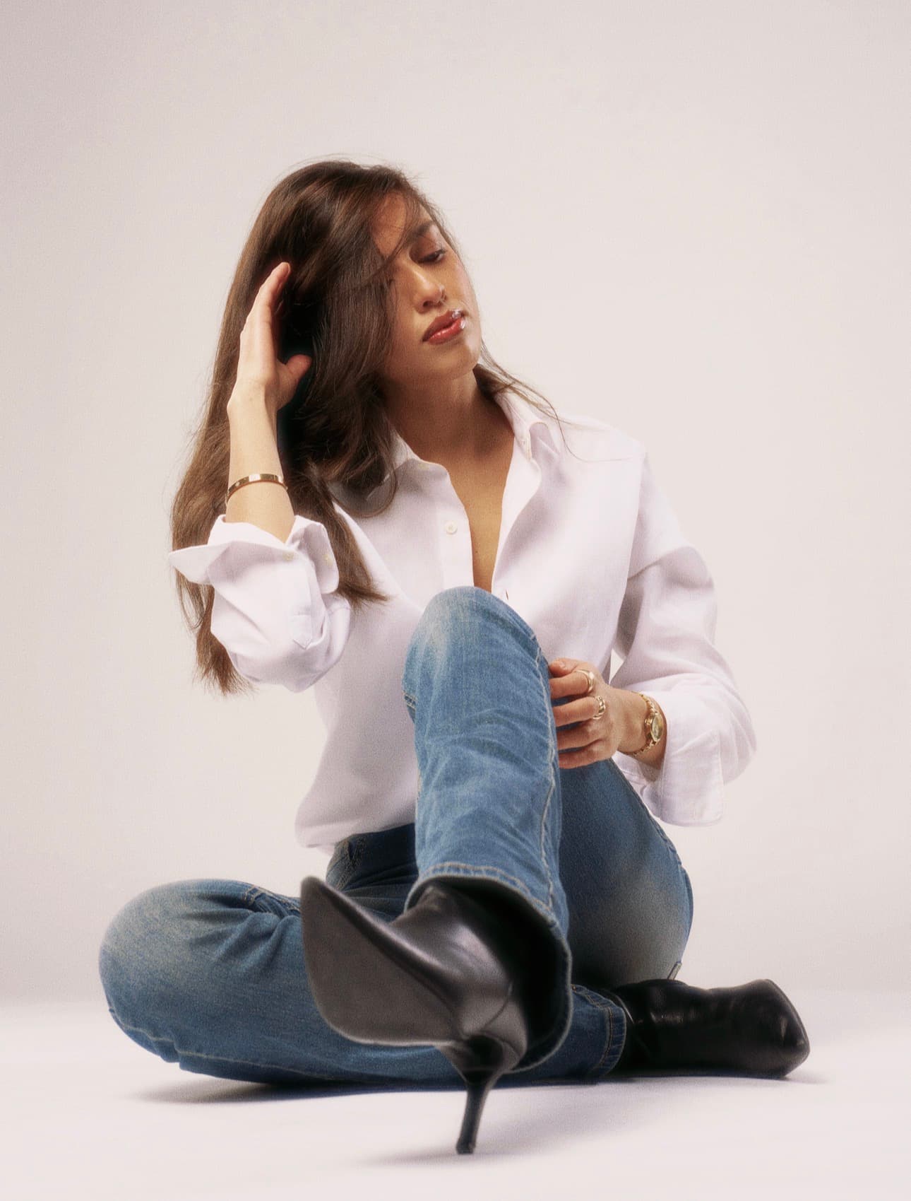 Woman in white shirt and jeans sits on floor, extending a black pointed-toe boot.