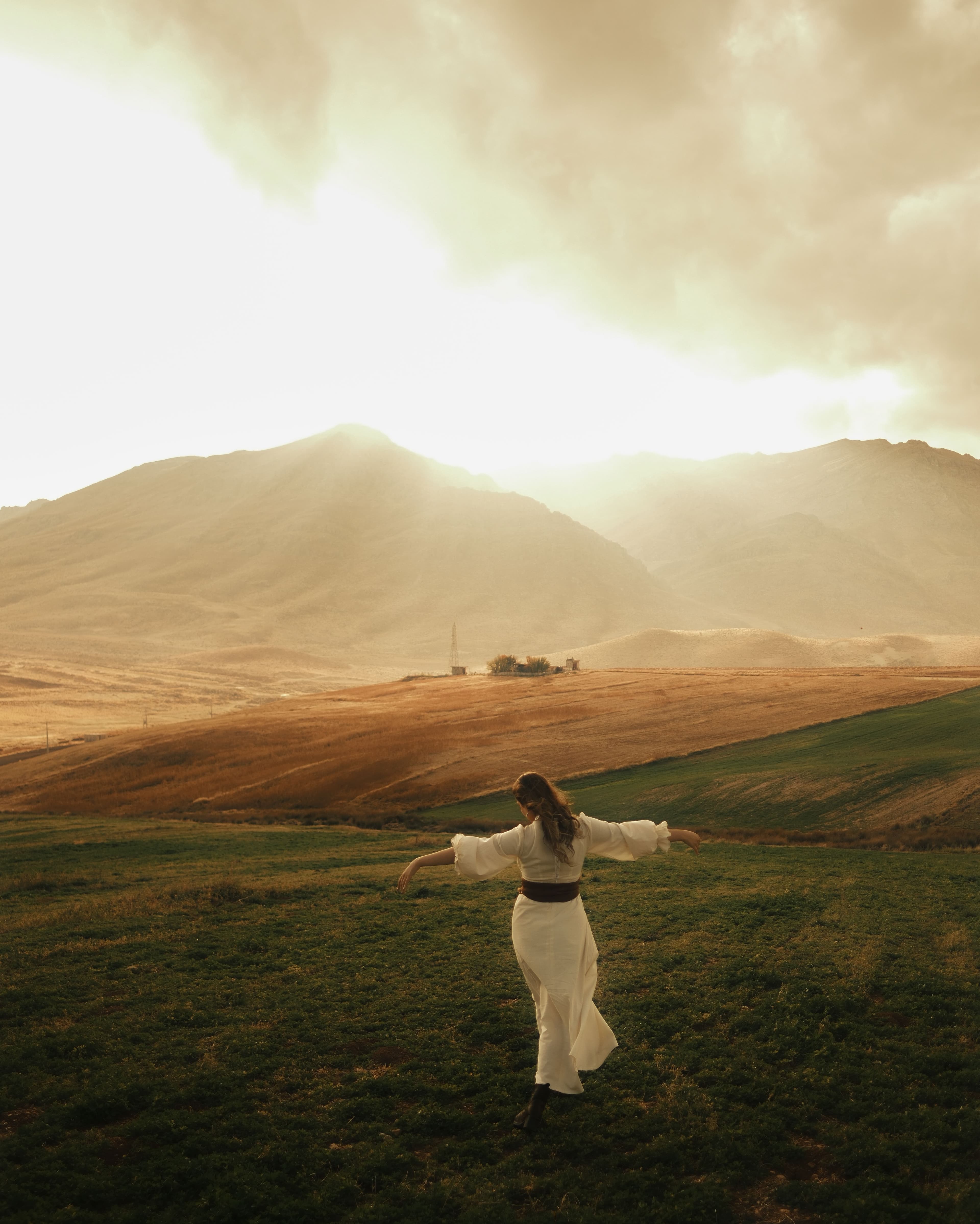Woman in white dress with arms outstretched in a field before hazy golden mountains.